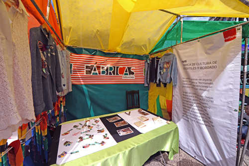 Piezas de joyería textil sobre mesa y prendas bordadas colgadas en percha al costado del stand